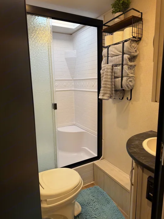This modern RV rental bathroom showcases a compact shower with black frame, toilet, vanity, and a wall shelf with towels.. Forest River Tracer 2022