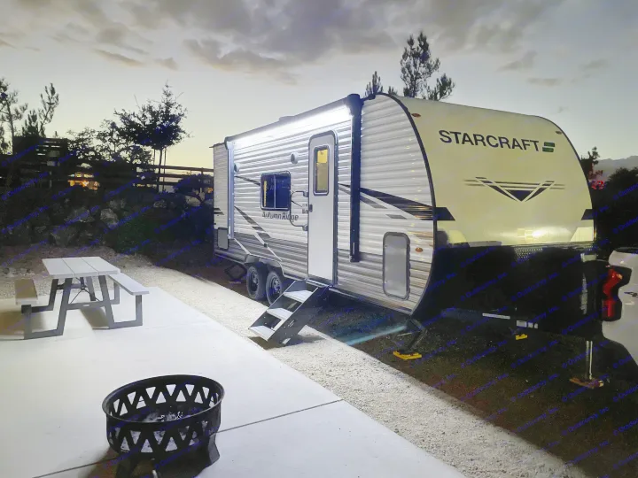 Brightly lit rv rental at a serene campsite with a picnic table & fire pit, under a dusky sky. Ideal for glamping.. Jayco Autumn Ridge 20MB 2023