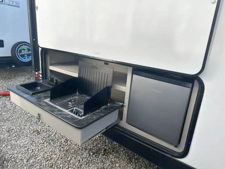 Outdoor kitchen of an RV rental: pull-out counter with sink, two-burner stove, and integrated compact fridge.. Forest River Tracer 2022