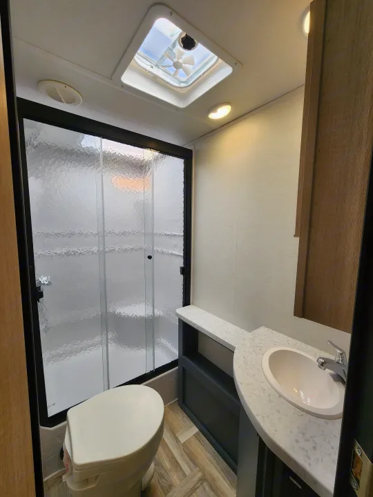 Inside an RV rental bathroom: compact design with textured glass shower, toilet, vanity with sink, skylight, and wood-look flooring.. primetime tracer 2024