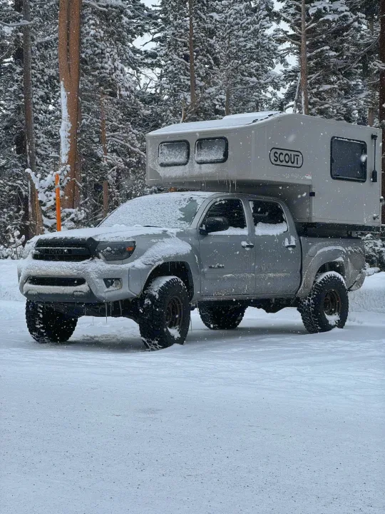 A grey Toyota truck with a "Scout" truck camper, covered in snow in a winter forest. Perfect for an RV rental adventure.. Adventurer 2015 Tacoma 2024