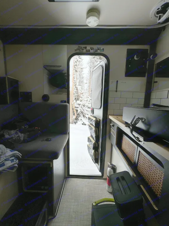 An RV rental interior, with an open door looking out onto a snow-covered forest. A kitchen counter and bench seating are visible.. Adventurer 2015 Tacoma 2024
