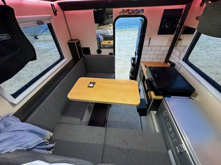 Cozy RV rental interior with a grey upholstered dining area, wooden table, and integrated kitchenette. Outside, a sandy shore.. Adventurer 2015 Tacoma 2024