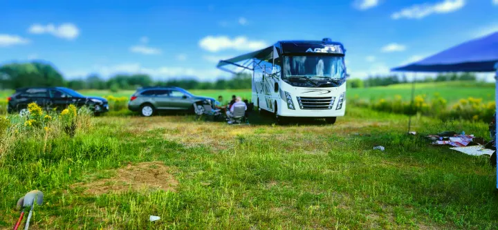 RV rental trip: People relax in chairs by a white motorhome with an awning, two cars, and a blue sky in a green field.. Thor A.C.E. 2020