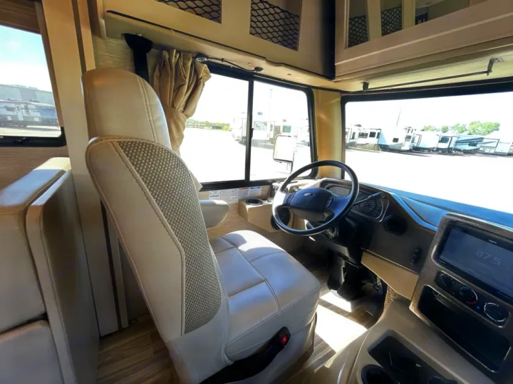 Inside an RV rental: view from the passenger side showing the driver's cockpit, seat, dashboard, and exterior RVs.. Thor A.C.E. 2020