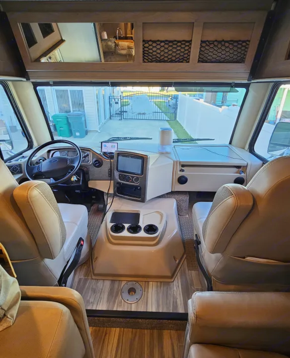 Inside an RV rental: The front driving area with beige seats, steering wheel, and dashboard with screens, showing the view outside.. Thor A.C.E. 2020