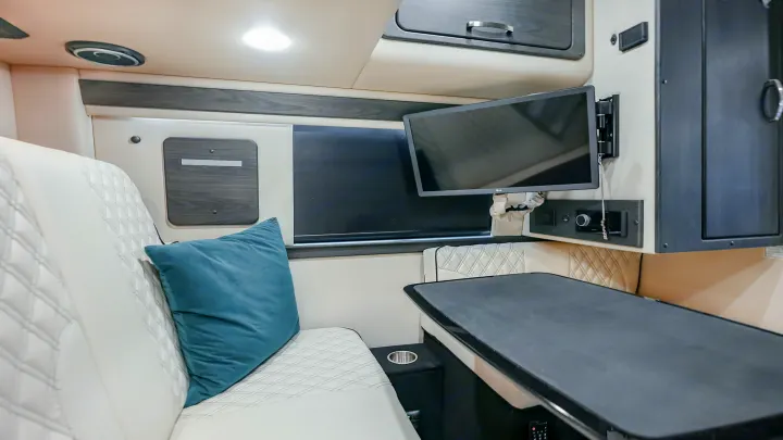 Spacious rv rental interior showing white quilted seating, a large table, and a mounted TV, ideal for comfortable travel or work.. American Coach Mercedes-Benz Sprinter 2023