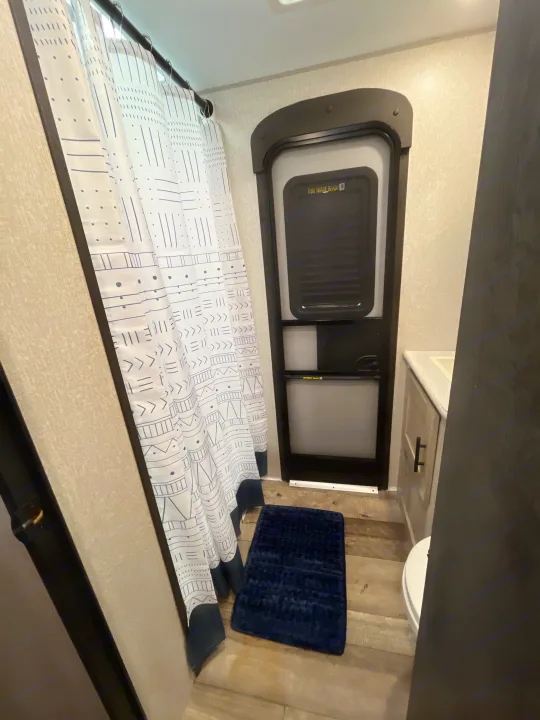 This RV rental photo shows a compact bathroom with a patterned shower curtain, blue mat, and entry door.. KZ Connect 2021