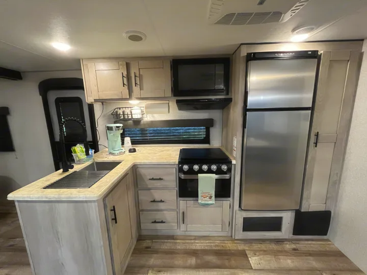 Bright, modern RV rental kitchen: light wood cabinets, stainless fridge, oven, microwave, sink, and wood-look floor.. KZ Connect 2021