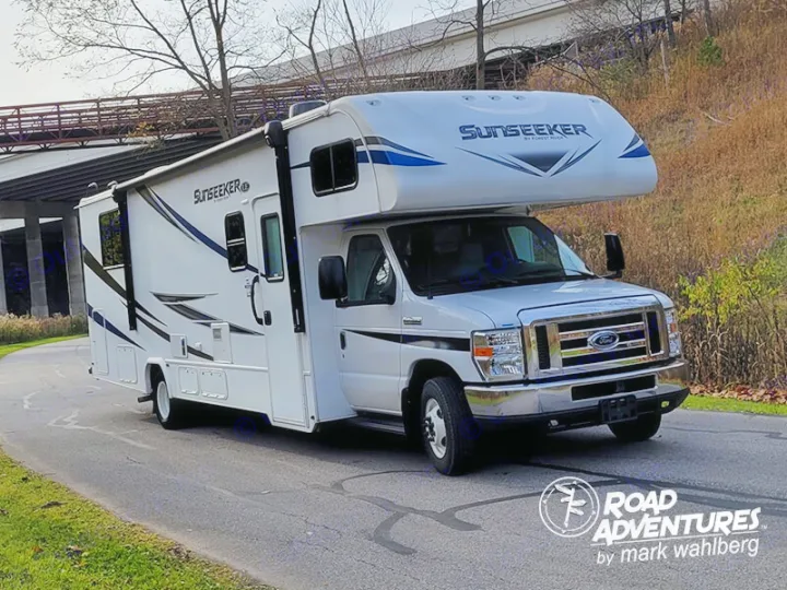 A white Sunseeker Class C RV rental, perfect for road adventures, is parked on a paved road with a bridge in the background.. Forest River Sunseeker 2027