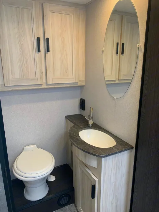Clean, modern RV rental bathroom featuring a toilet, vanity with sink, oval mirror, and light wood grain cabinets.. Forest River Sunseeker 2027
