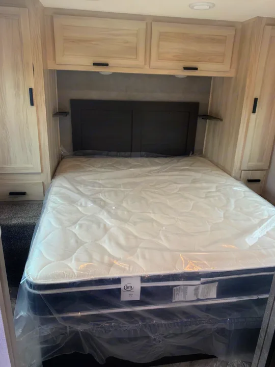 RV rental bedroom with a new Serta queen mattress (in plastic), dark headboard, and light wood built-in storage.. Forest River Sunseeker 2027