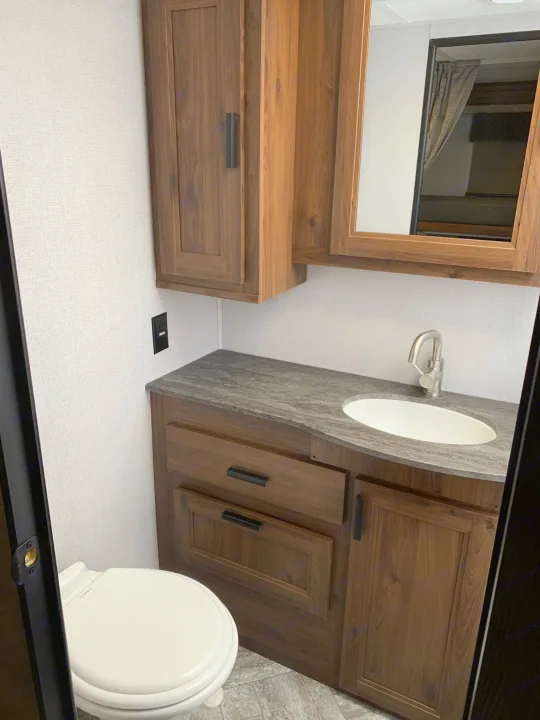 This RV rental features a compact bathroom with a toilet, wood vanity, sink, and overhead mirrored cabinets.. Forest River Sunseeker 2026