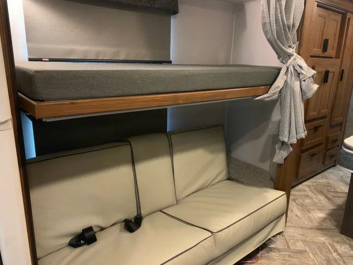 RV rental interior: a grey bunk bed over a light sofa with seatbelts, next to wood cabinets.. Forest River Sunseeker 2026