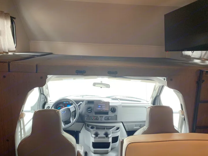 Inside this RV rental: front cab with dashboard, steering wheel, seats, and a cozy overhead bunk with TV.. Forest River Sunseeker 2026