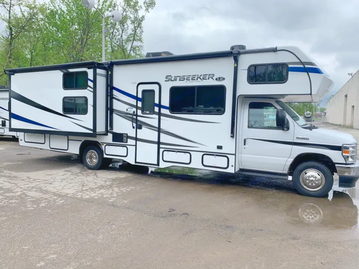 A white Sunseeker RV rental with blue/black graphics is parked on a wet asphalt lot. This Class C motorhome has an extended slide-out.. Forest River Sunseeker 2026