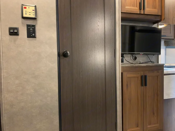 An RV rental interior: wall with controls (thermostat, slide out), a wooden door, and a mounted TV above wood cabinets.. Forest River Sunseeker 2026