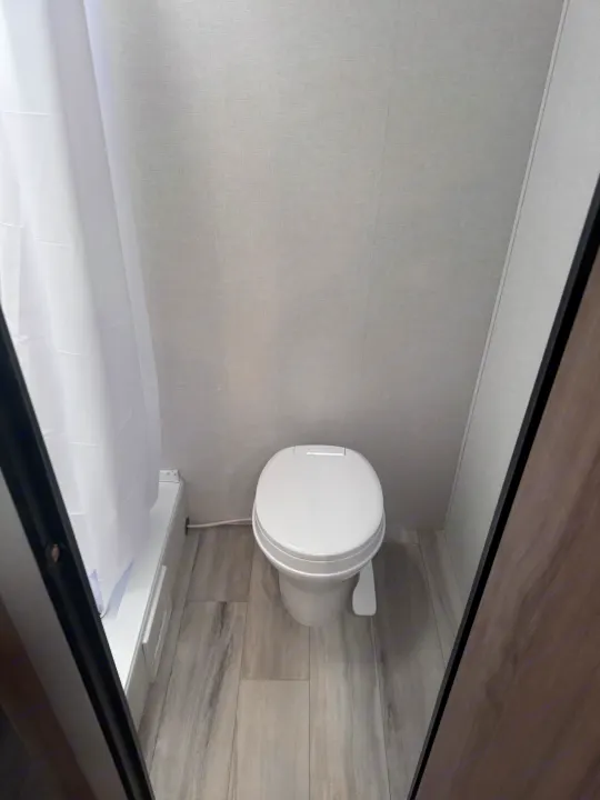 Compact RV rental bathroom with a white toilet, grey wood-look floor, and a shower area with a white curtain.. Keystone Coleman 17B 2025