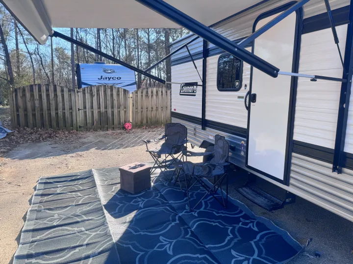 Sunny RV rental campsite with a cozy outdoor living area: extended awning, two chairs, and a patterned mat.. Keystone Coleman 17B 2025