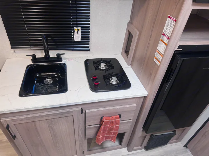 RV rental kitchen area: compact black sink, two-burner stove, light counter, wood cabinets, and built-in refrigerator.. Keystone Coleman 17B 2025