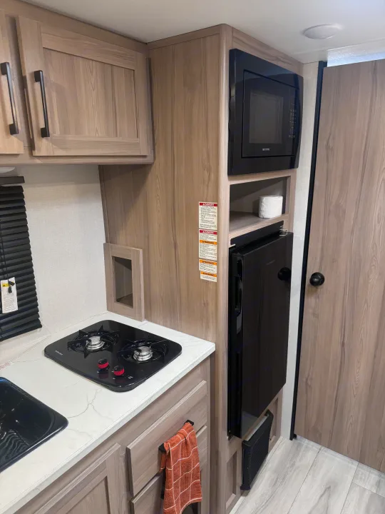 Compact RV rental kitchen featuring a two-burner stove, microwave, and built-in refrigerator with wood-look cabinets.. Keystone Coleman 17B 2025