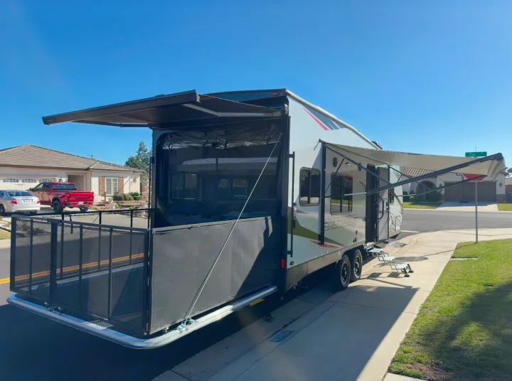 This RV rental, a toy hauler, is set up with a rear patio deck and extended awnings on a sunny suburban street.. Forest River Shockwave 2021