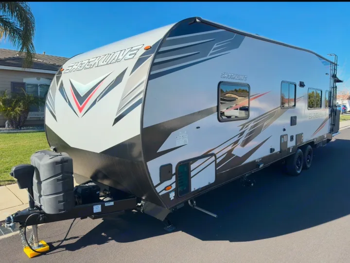 This white and black Shockwave travel trailer, perfect for an RV rental, features bold graphics & dual axles, parked on a sunny street.. Forest River Shockwave 2021