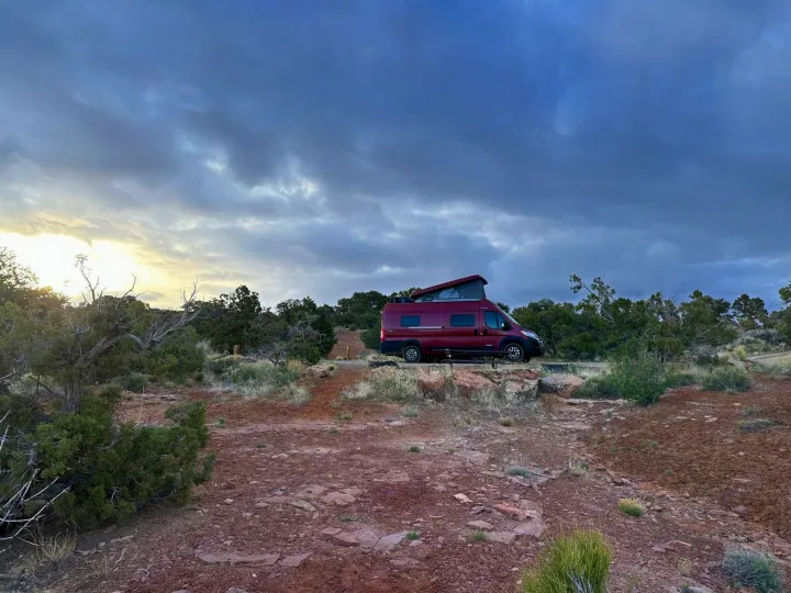 Enjoy nature with an RV rental! A red pop-top camper van is parked on rocky desert land under a dramatic cloudy sky.. Winnebago Solis 2023
