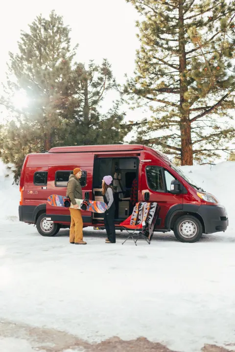 Snowy RV rental trip: Man with snowboard, woman by red camper van, chair, blanket in winter scene.. Winnebago Solis 2022