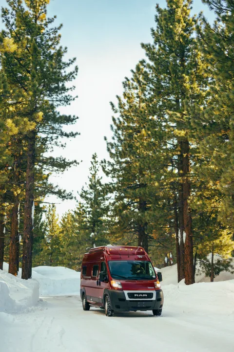 A red RV rental van drives on a snowy road through a pine forest, perfect for winter adventures.. Winnebago Solis 2022