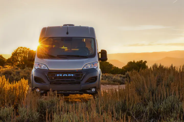 Light grey Ram ProMaster RV rental parked in golden desert brush at sunset, with sun rays peeking over the horizon.. Dodge Ram 1500 2024