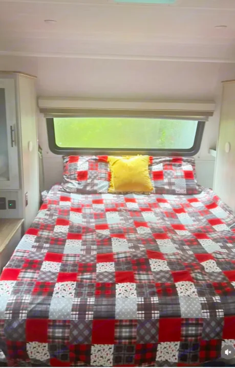 Inside a comfortable RV rental: a bed with a red plaid comforter, yellow accent pillow, and nature view from the window.. Forest River IBEXMBS 2023