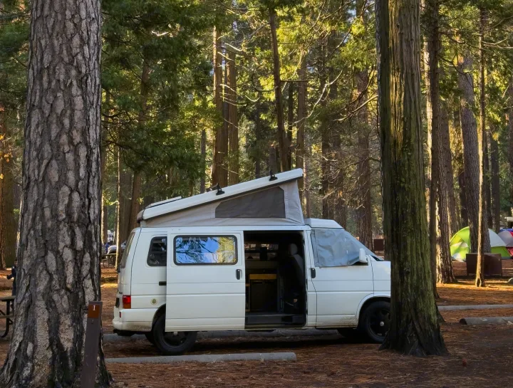 An RV rental pop-top camper van at a sunny forest campsite with its side door open, surrounded by tall trees and tents.. Winnebago Eurovan Camper Eurovan Camper 1999