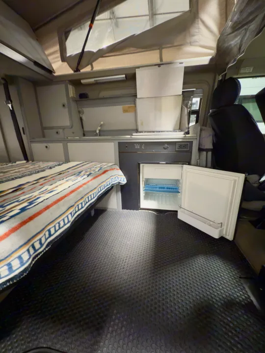Inside an RV rental: a bed with patterned blanket, kitchen, and open mini-fridge. Pop-top roof visible.. Winnebago Eurovan Camper Eurovan Camper 1999