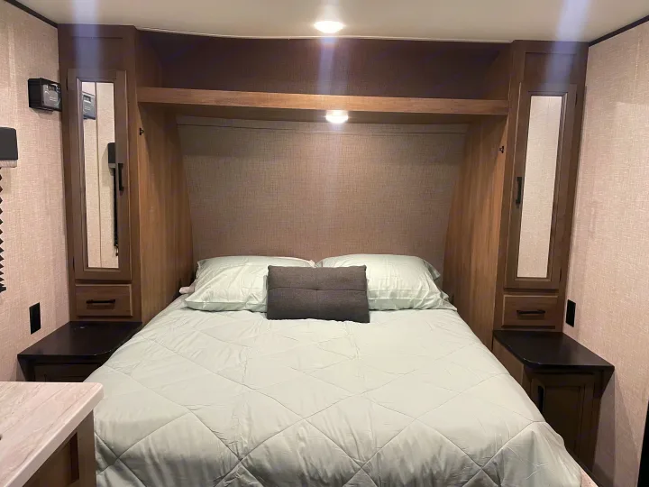 An RV rental bedroom features a cozy bed with light green bedding, flanked by mirrored storage cabinets, nightstands, and overhead lighting.. Jayco Autumn Ridge 20MB 2023