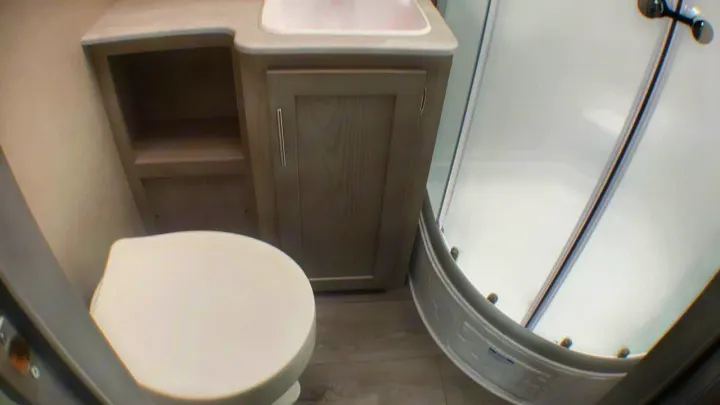 A compact RV rental bathroom with a white toilet, light wood vanity, sink, open shelving, and a curved glass shower.. Coachmen Catalina 2021