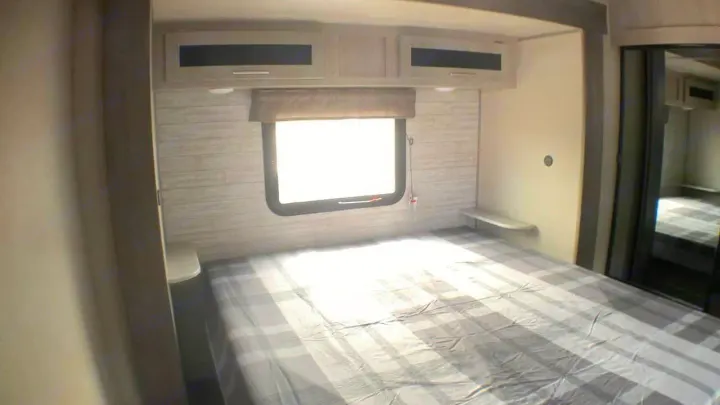 An inviting RV rental bedroom featuring a plaid bed, window, overhead cabinets, and a mirrored closet on the right.. Coachmen Catalina 2021
