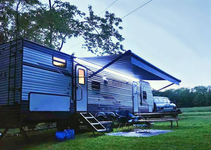 An RV rental illuminates an evening campsite with its extended awning, chairs, and picnic table on the grass.. Palomino Puma 2024