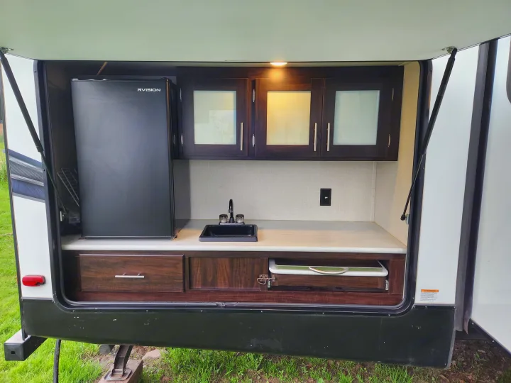 RV rental outdoor kitchen with a black fridge, sink, dark wood cabinets, light countertop, and pull-out tray.. Prime Time Lacrosse 2019