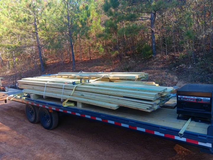 Heavy-duty trailer with strapped stacks of treated lumber on a dirt path in woods. Good for home building or an RV rental project.. Blazer Deck over / Flatbed 2023