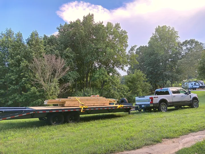 White truck hauls lumber on a flatbed trailer through a grassy field. RVs in the background suggest an RV rental area.. Blazer Deck over / Flatbed 2023