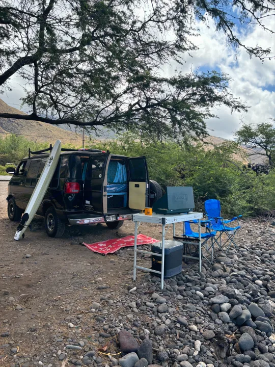 Explore with an RV rental! Camper van with surfboard, portable stove, and chairs set up on a rocky shore under a large tree.. Dodge 1500 1999