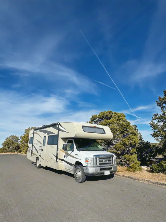 White and beige RV rental parked on an asphalt road, surrounded by green trees, under a blue sky with multiple crisscrossing contrails.. Coachmen Leprechaun 2019