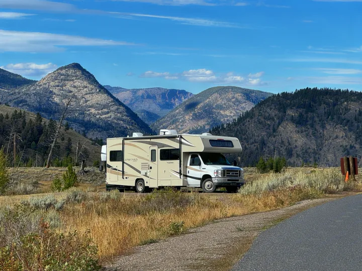 A beige Class C rv rental is parked on a dirt roadside with scenic mountains and a blue sky. Ideal for outdoor adventures.. Coachmen Leprechaun 2019