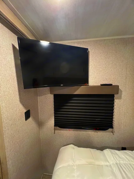 Inside an **rv rental**: a cozy bedroom with a wall-mounted TV, window blind, and comfortable white bedding.. East to West Silver Lake 2022