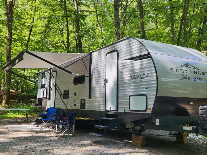 A well-equipped RV rental travel trailer with an extended awning and chairs at a shaded, wooded campground for camping.. East to West Silver Lake 2022