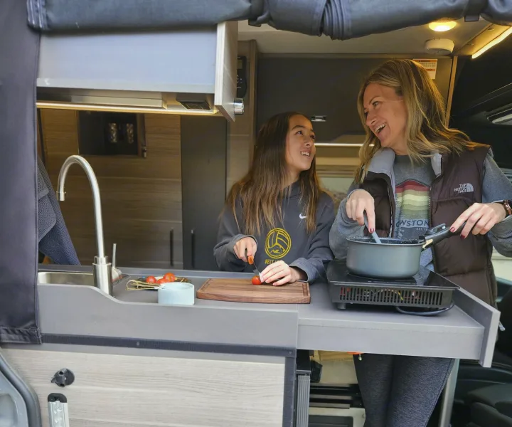 Happy women cooking in their RV rental kitchen. One stirs a pot on the burner, the other preps tomatoes by the sink.. Entegra Coach Launch 2023