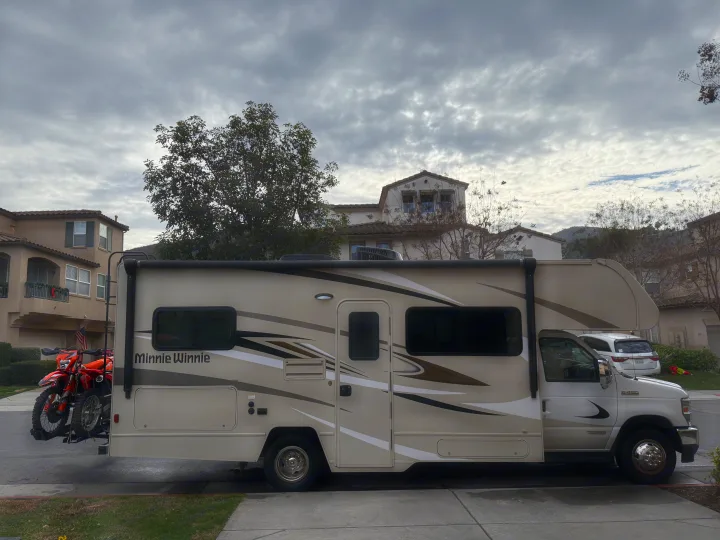 A beige Minnie Winnie RV rental with two orange dirt bikes mounted on the back, parked on a residential street.. Winnebago Minnie Winnie 2020