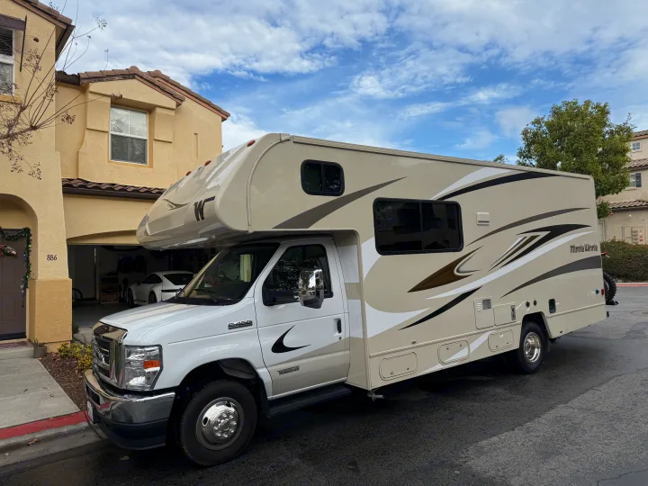 White and beige Winnebago RV rental parked in front of a house with a visible garage and car, under a cloudy sky.. Winnebago Minnie Winnie 2020
