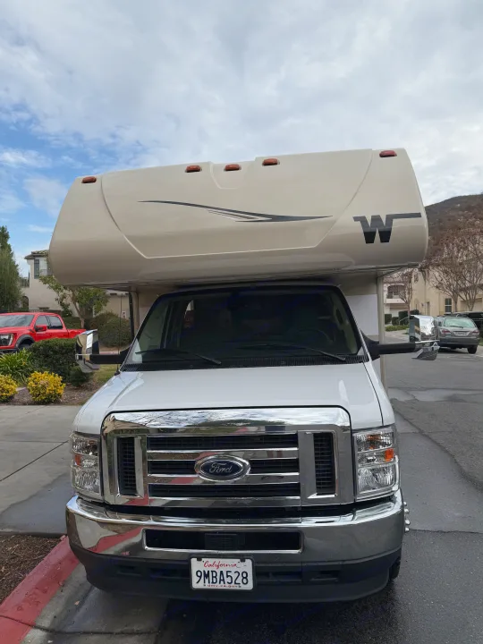 Front view of a white Ford RV rental, featuring a chrome grill and a large 'W' logo. Parked in a residential area.. Winnebago Minnie Winnie 2020